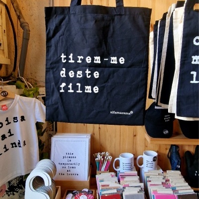 Saco de tecido azul escuro com texto branco pendurado e vários produtos de papelaria e acessórios em mesa de madeira
