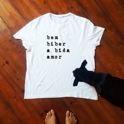 T-shirt branca com texto em preto deitada no chão de madeira, gato preto ao lado e pés humanos visíveis.