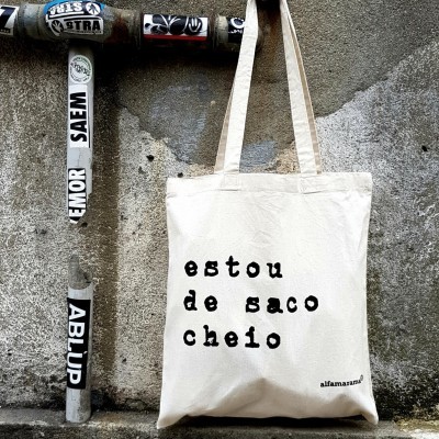 Saco de tecido cru com texto 'estou de saco cheio' pendurado numa parede de cimento rugosa