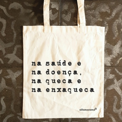 Saco de tecido branco com texto preto e alças sobre chão cinzento com padrão abstrato