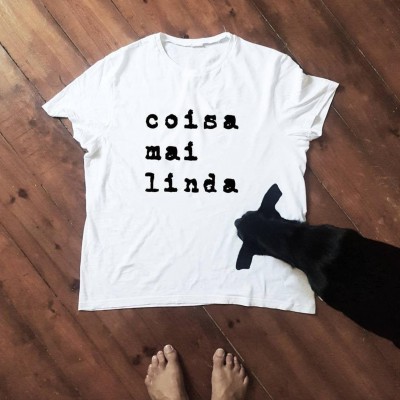 T-shirt branca com texto 'coisa mai linda' no chão de madeira com cabeça de cabra preta ao lado