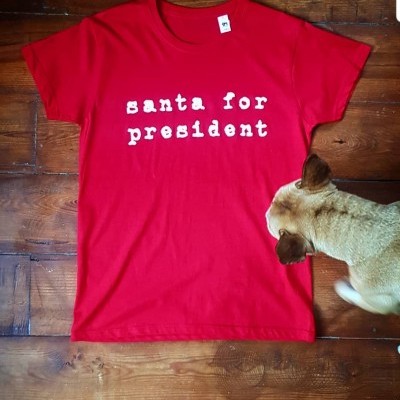Camiseta vermelha com texto branco 'santa for president' sobre chão de madeira e cão ao lado.