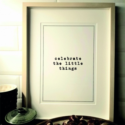 Quadro com moldura dourada claro e texto em letras pretas, junto a tigela de castanhas e cerâmica sobre superfície, fundo de azulejos brancos.