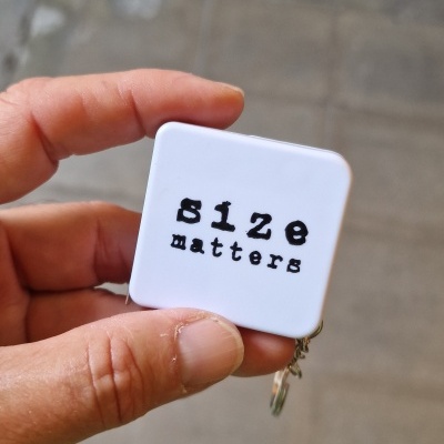 Medidor de fita métrica branco com texto 'size matters' segurado por mão.