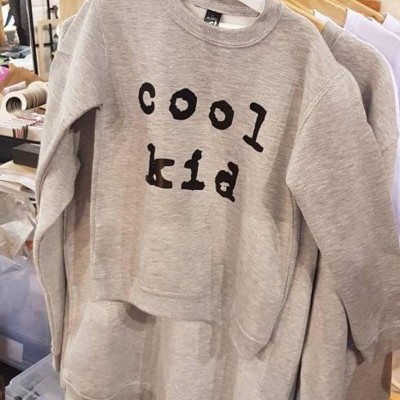 Camisola cinzenta de criança com texto preto 'cool kid' pendurada num cabide branco numa loja