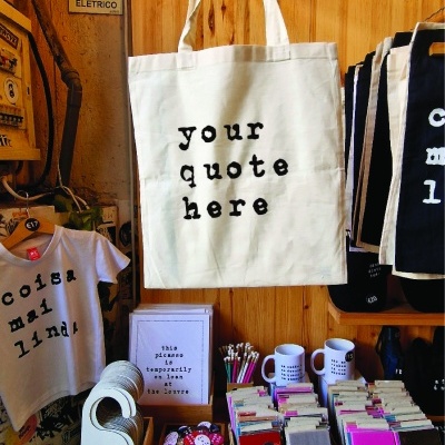 Sacola de tecido bege com texto preto, t-shirt branca, artigos variados em mesa de madeira