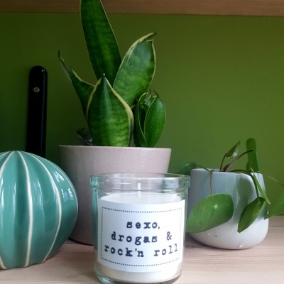 Vela em vidro com texto e duas plantas em vasos sobre superfície de madeira