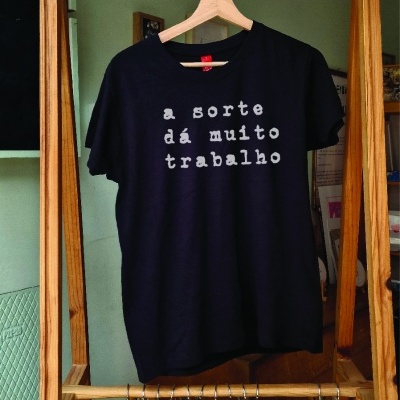 T-shirt preta com texto em letras brancas pendurada em estrutura de madeira com sacos de tecido brancos.