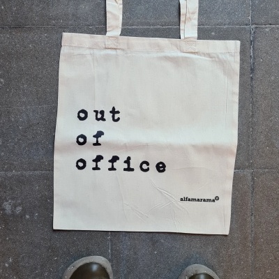 Saco tote de pano bege claro com texto preto 'out of office' colocado no chão