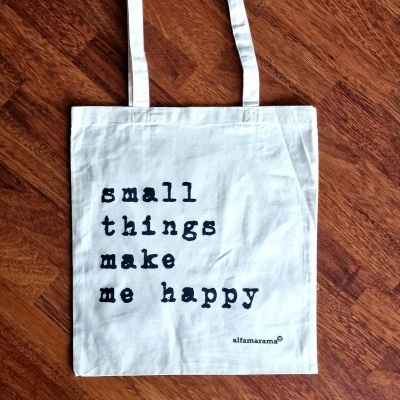 Saco de tecido branco com texto preto e alças sobre piso de madeira