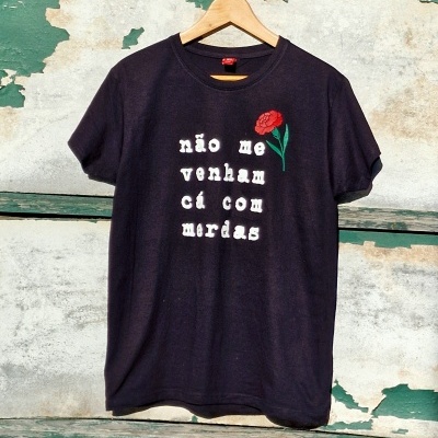 T-shirt preta com texto branco e flor vermelha pendurada em cabide contra parede velha verde e branca
