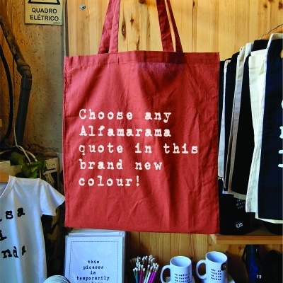 Saco vermelho com texto branco pendurado em suporte de madeira num espaço com parede branca texturada e vários artigos de papelaria e canecas ao redor.