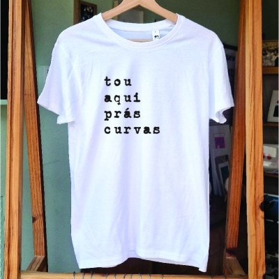 T-shirt branca com texto preto pendurada numa estrutura de madeira com calças em cabides