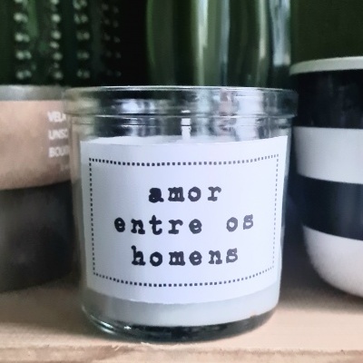 Vela em vidro com etiqueta de texto e vasos decorativos atrás numa prateleira de madeira