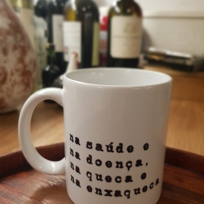 Caneca branca com texto em português colocada num tabuleiro de madeira com garrafas ao fundo