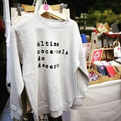 Sweatshirt cinza claro com texto preto 'última coca-cola do deserto' à venda num mercado ao ar livre