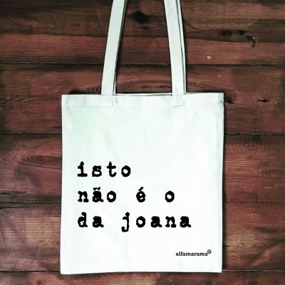 Saco de tecido branco com texto impresso preto pendurado numa parede de madeira.