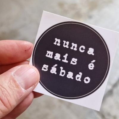 Autocolante quadrado com texto em branco num círculo preto, segurado por uma mão.