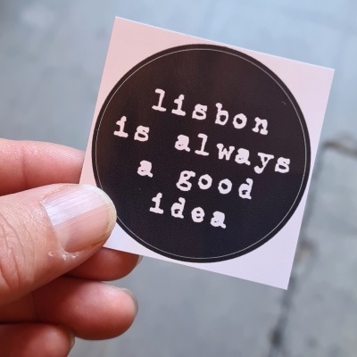 Adesivo quadrado branco com círculo preto e texto branco 'lisbon is always a good idea'