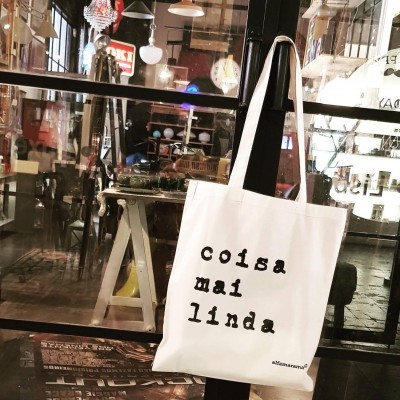 Saco de tecido branco com texto preto 'coisa mai linda' pendurado em loja