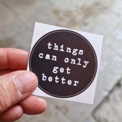 Autocolante com texto 'things can only get better' numa mão, fundo de chão e parede de pedra.