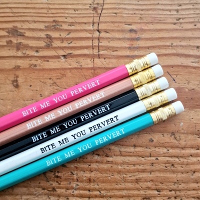 Seis lápis coloridos alinhados com texto 'BITE ME YOU PERVERT' sobre mesa de madeira