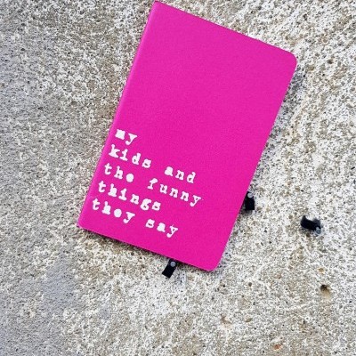 Caderno rosa com texto branco 'my kids and the funny things they say' numa superfície cinzenta rugosa