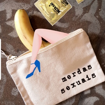 Bolsa de tecido bege com texto 'merdas sexuais', banana e preservativos em embalagens douradas sobre superfície marmoreada cinzenta