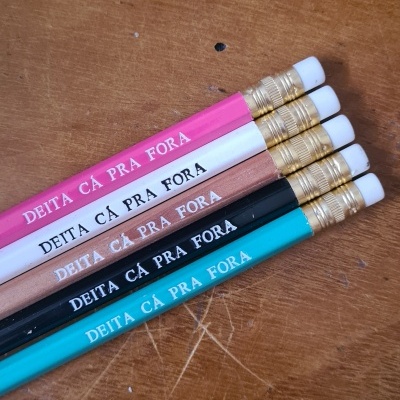 Cinco lápis coloridos com frase 'DEITA CÁ PRA FORA' sobre mesa de madeira