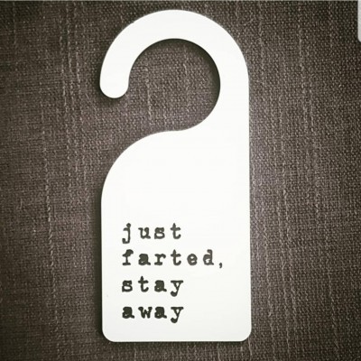 Pendurador de porta branco com texto preto 'just farted, stay away' sobre superfície castanha