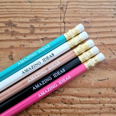 Cinco lápis coloridos com borracha e texto 'AMAZING IDEAS' sobre madeira rústica