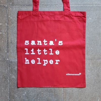 Saco reutilizável vermelho com texto branco 'santa's little helper' em tecido, com alças longas, sobre chão de pedra