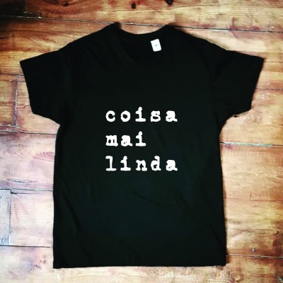 T-shirt preta com texto branco 'coisa mai linda' sobre chão de madeira
