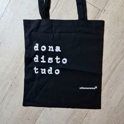 Saco de pano preto com texto branco e alças longas sobre piso de madeira clara