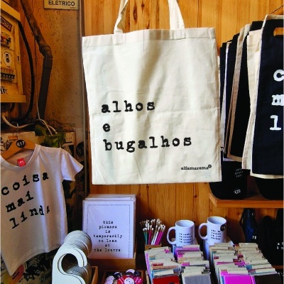 Saco de tecido branco com texto, t-shirt branca com texto e vários produtos artesanais em bancada de madeira.