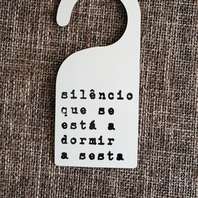 Porta para pendurar branco com texto preto 'silêncio que se está a dormir a sesta' sobre fundo têxtil castanho