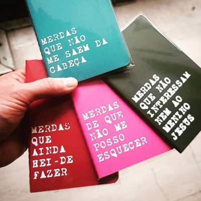Quatro cadernos coloridos com texto branco em maiúsculas, segurados por uma mão.