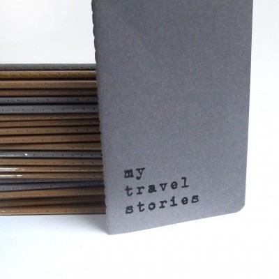 Caderno cinzento com texto 'my travel stories' em frente a pilha de cadernos