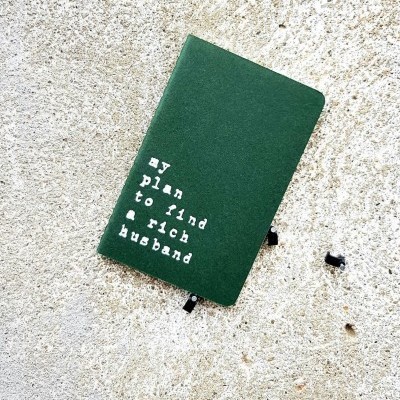 Caderno verde com texto branco 'my plan to find a rich husband'