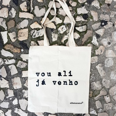 Saco de tecido crú com texto preto pendurado numa parede de pedra