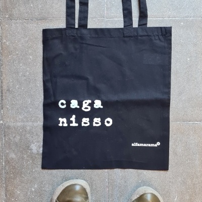 Saco de tecido preto com texto branco colocado no chão com calças cor de salmão e sapatilhas verdes visíveis