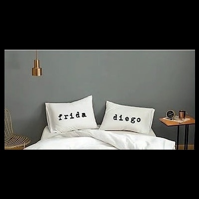Almofadas brancas com nomes em preto numa cama branca e ambiente cinza