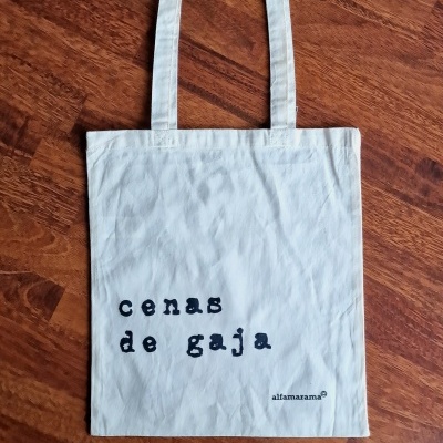 Sacola branca com texto preto 'cenas de gaja' sobre chão de madeira.