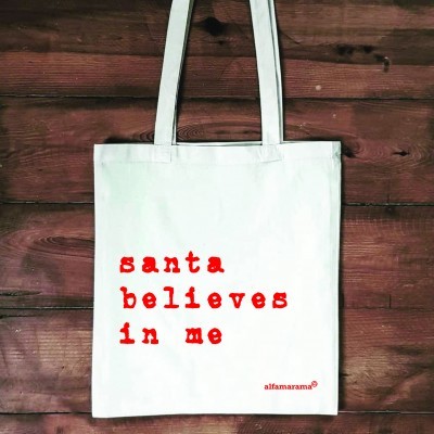 Saco branco com texto vermelho 'santa believes in me' e alças finas, pendurado numa parede de madeira.