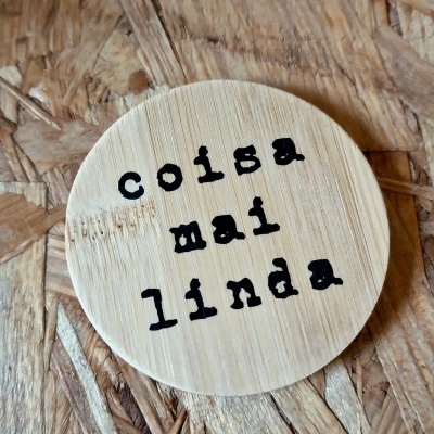 Placa redonda de madeira clara com texto 'coisa mal linda' sobre superfície de madeira aglomerada