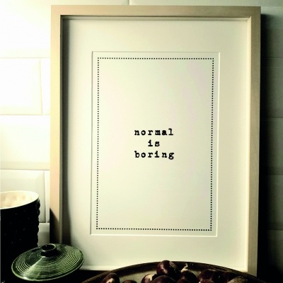 Quadro com texto 'normal is boring' em moldura bege clara sobre superfície com tigela verde e bandeja com frutos marrons.
