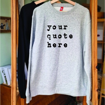 Sweatshirt cinzenta com texto preto pendurada em cabide de madeira numa estrutura de madeira.