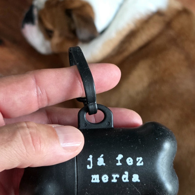 Recipiente preto para sacos de dejetos em formato de osso com texto branco e cão desfocado ao fundo.