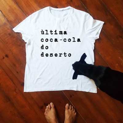 Camisola branca com texto em preto, chão de madeira, cão preto e pés humanos