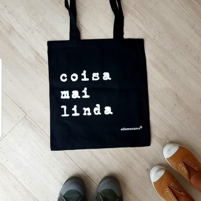 Saco preto com texto branco 'coisa mai linda' junto de duas sapatilhas no chão